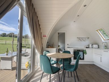 A kitchen and dining area with a round wooden table and teal chairs a toaster and oven in a white interior at Maple in Wootton Fitzpaine near Charmouth