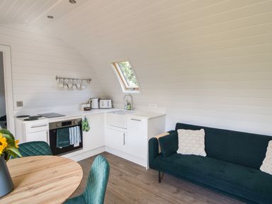 A kitchen with white cabinets and appliances next to a dark green sofa with cushions at Maple in Wootton Fitzpaine near Charmouth