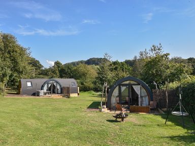 Two pods with outdoor seating on a grassy lawn with trees and hills in the background at Maple Wootton Fitzpaine near Charmouth