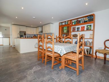 A dining area with wooden chairs and a table with a polka dot tablecloth next to a kitchen with white cabinets and a wooden bookshelf filled with books at The Orchard in Cardinham