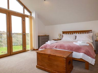 A bedroom with a wooden bed a pink blanket bedside tables lamps a wooden chest and large windows at The Orchard in Cardinham