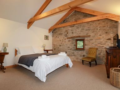 A bedroom with a bed with white and grey bedding two bedside tables with lamps a stone wall wooden ceiling beams a yellow chair a wooden dresser with a TV and a wicker basket at The Orchard in Cardinham