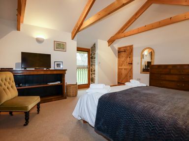 A bedroom with a bed covered with a dark blanket a TV on a wooden shelf a wooden dresser a chair and wooden beams on the ceiling at The Orchard in Cardinham