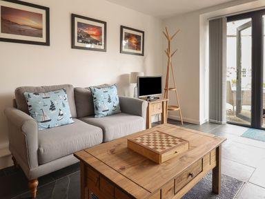 A living room with a gray sofa and blue cushions with sailboat patterns a wooden coffee table with a chessboard a small wooden side table with a lamp and a TV a wooden coat rack and a glass door leading outside at The Hideaway in Uplyme near Lyme Regis