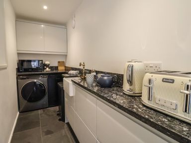 A narrow kitchen with a black washing machine a microwave toaster kettle teapot and a brass faucet at The Hideaway in Uplyme near Lyme Regis