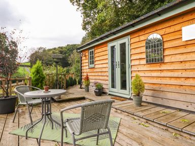 An outdoor wooden deck with a metal table and two chairs near a wooden building with green double doors and potted plants at Chateau Petit in Trefeglwys