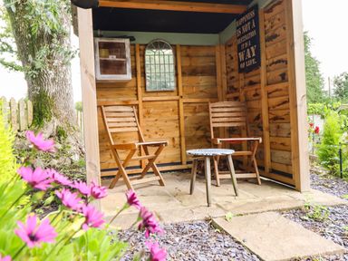 A wooden garden shelter with two wooden chairs and a small round table surrounded by flowers at Chateau Petit in Trefeglwys