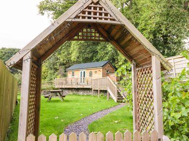 A wooden garden archway leading to a small wooden cabin with a deck and stairs in a grassy backyard at Chateau Petit in Trefeglwys