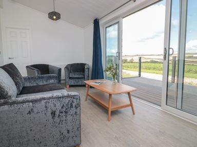 A living room with a sofa, two armchairs, a wooden coffee table with flowers, and large sliding glass doors opening to a deck at Buttercup Lodge in Norton near Dartmouth