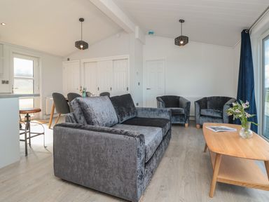 A living room with a grey sofa two armchairs a wooden coffee table with flowers and magazines and a dining table with chairs at Buttercup Lodge Norton near Dartmouth