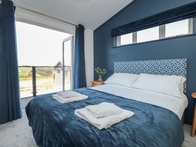 A bedroom with a blue bedspread white pillows a flower vase on a wooden nightstand and a door leading outside at Buttercup Lodge in Norton near Dartmouth
