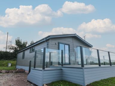 A small gray house with glass railings on a raised deck surrounded by grass and a cloudy sky at Honeysuckle Lodge in Norton near Dartmouth