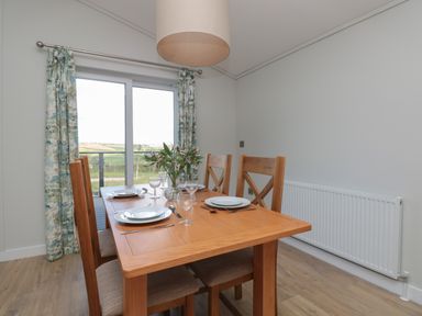 A dining table with four wooden chairs set with plates and glasses next to a sliding glass door with patterned curtains at Honeysuckle Lodge in Norton near Dartmouth