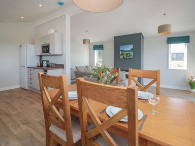 A dining area with a wooden table and chairs set with plates and glasses next to a kitchen with white cabinets and a white refrigerator at Honeysuckle Lodge in Norton near Dartmouth