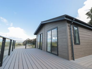 A wooden deck with glass railing attached to a small building with sliding glass doors at Primrose Lodge in Norton near Dartmouth