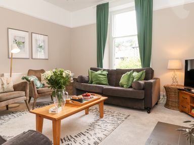 A living room with a brown sofa and green cushions a wooden coffee table with a vase of flowers and a tray two armchairs a floor lamp and framed pictures on a wall at Church View in Grange-Over-Sands