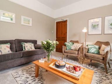 A living room with a gray sofa, two armchairs, a wooden coffee table with a vase of flowers and a tray of snacks at Church View in Grange-Over-Sands