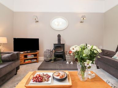 A living room with a wood stove a television on a wooden stand and a coffee table with a vase of flowers and a tray of pastries at Church View in Grange-Over-Sands