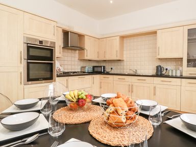 A kitchen with wooden cabinets and built in oven a dining table set with plates bowls glasses and baskets of fruit and bread at Church View in Grange-Over-Sands