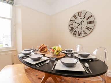 A dining room with a black oval table set with bowls glasses cutlery a basket of croissants and a bowl of fruit with clear chairs and a large wall clock at Church View in Grange-Over-Sands