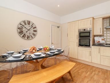 A dining area with a black oval table set with plates bowls and glasses clear chairs a wooden bench a wall clock and light wood kitchen cabinets at Church View in Grange-Over-Sands