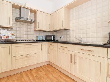 A kitchen with wooden cabinets a black countertop a gas stove a blue toaster a microwave and a sink at Church View in Grange-Over-Sands