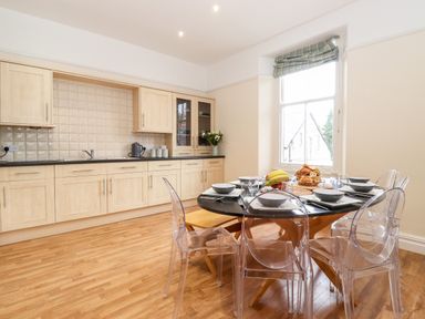 A kitchen with wooden cabinets a round dining table set with bowls and glasses and transparent chairs at Church View in Grange-Over-Sands