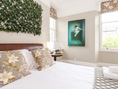 A bedroom with a bed with floral pillows green wall decoration artwork on the wall and two windows at Church View in Grange-Over-Sands
