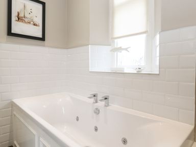 A bathroom with a white bathtub white tiled walls a window with a blind and a framed picture at Church View in Grange-Over-Sands
