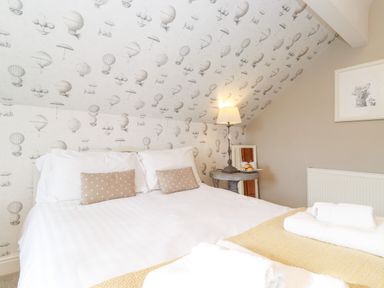 A bedroom with a bed with white bedding and beige pillows next to a round table with a lamp and croissants at Church View in Grange-Over-Sands