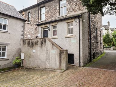 An exterior view of a stone and stucco building with stairs leading to a black door and a paved parking area at Church View in Grange-Over-Sands