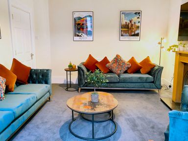 A living room with blue sofas and orange cushions a round wooden coffee table with a plant and wall art at Cranford House in Keswick