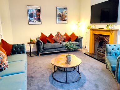 A living room with blue sofas orange cushions a round wooden coffee table a fireplace and a wall-mounted TV at Cranford House in Keswick