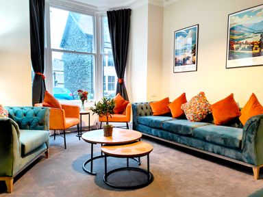 A living room with green sofas orange chairs and round wooden tables at Cranford House in Keswick