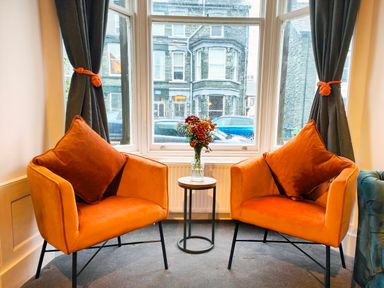 A seating area with two orange armchairs and a small round table with flowers in a vase by a window with grey curtains at Cranford House in Keswick