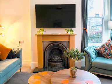A living room with a fireplace a wall mounted television green sofas a wooden coffee table and windows at Cranford House in Keswick