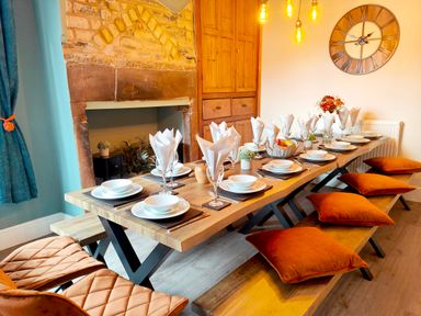 A dining room with a long wooden table set for a meal with plates napkins and glasses with orange cushions on benches and chairs at Cranford House in Keswick