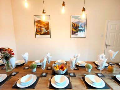 A dining table set with plates bowls utensils glasses with napkins and a fruit basket with wall art at Cranford House in Keswick