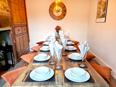 A dining room with a long wooden table set with white plates bowls utensils folded napkins glasses and orange cushions on the floor at Cranford House Keswick