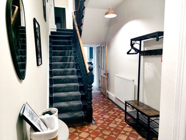 A hallway with stairs carpeted in dark fabric along with a wooden handrail a round mirror on the left wall a table with baskets underneath a light fixture hanging from the ceiling a coat rack with hangers and a radiator on the right wall at Cranford House in Keswick