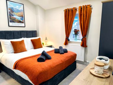 A bedroom with a bed with orange blanket and pillows a window with orange curtains and a bedside table with lamp at Cranford House Keswick