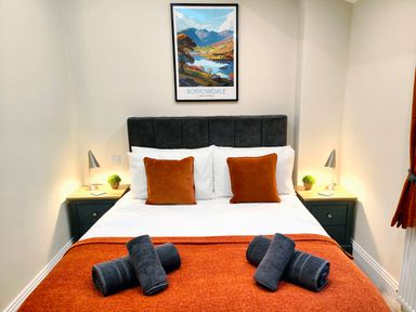 A bedroom with a bed with orange cushions and blanket two rolled towels on the bed two bedside tables with lamps and small plants borrowing poster on the wall at Cranford House in Keswick