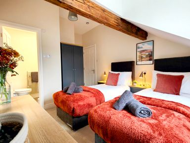 A bedroom with two single beds with orange throws and gray towels a wooden beam on the ceiling and a doorway to a bathroom at Cranford House in Keswick