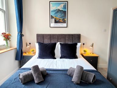 A bedroom with a bed with dark headboard two black pillows bedside tables with lamps and plants rolled towels on a blue blanket at Cranford House Keswick