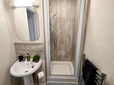 A small bathroom with a white pedestal sink a mirror above it a shower with a glass door and a towel rack with a black towel