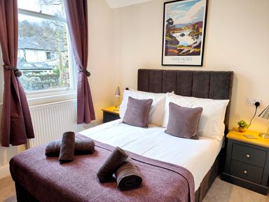 A bedroom with a double bed with purple pillows and blankets next to a window with purple curtains at Cranford House Keswick