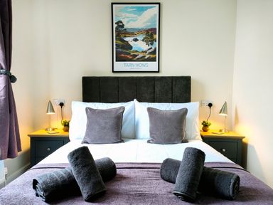 A bedroom with a bed pillows rolled towels bedside tables lamps and a framed landscape picture on the wall at Cranford House in Keswick