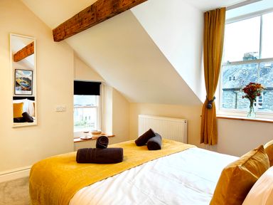A bedroom with a bed covered with a yellow and white bedspread and dark towels placed on it and windows with yellow curtains at Cranford House in Keswick