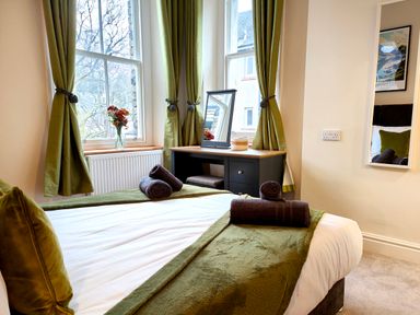 A bedroom with a bed with green blankets and pillows next to windows with green curtains and a desk with a mirror and flowers at Cranford House in Keswick
