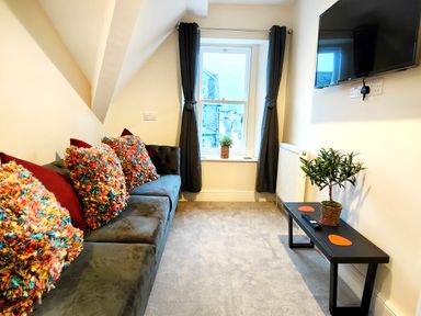 A small living room with a gray sofa with colorful pillows a window with dark curtains a wall-mounted TV and a black coffee table with a plant and remote controls at Cranford House in Keswick